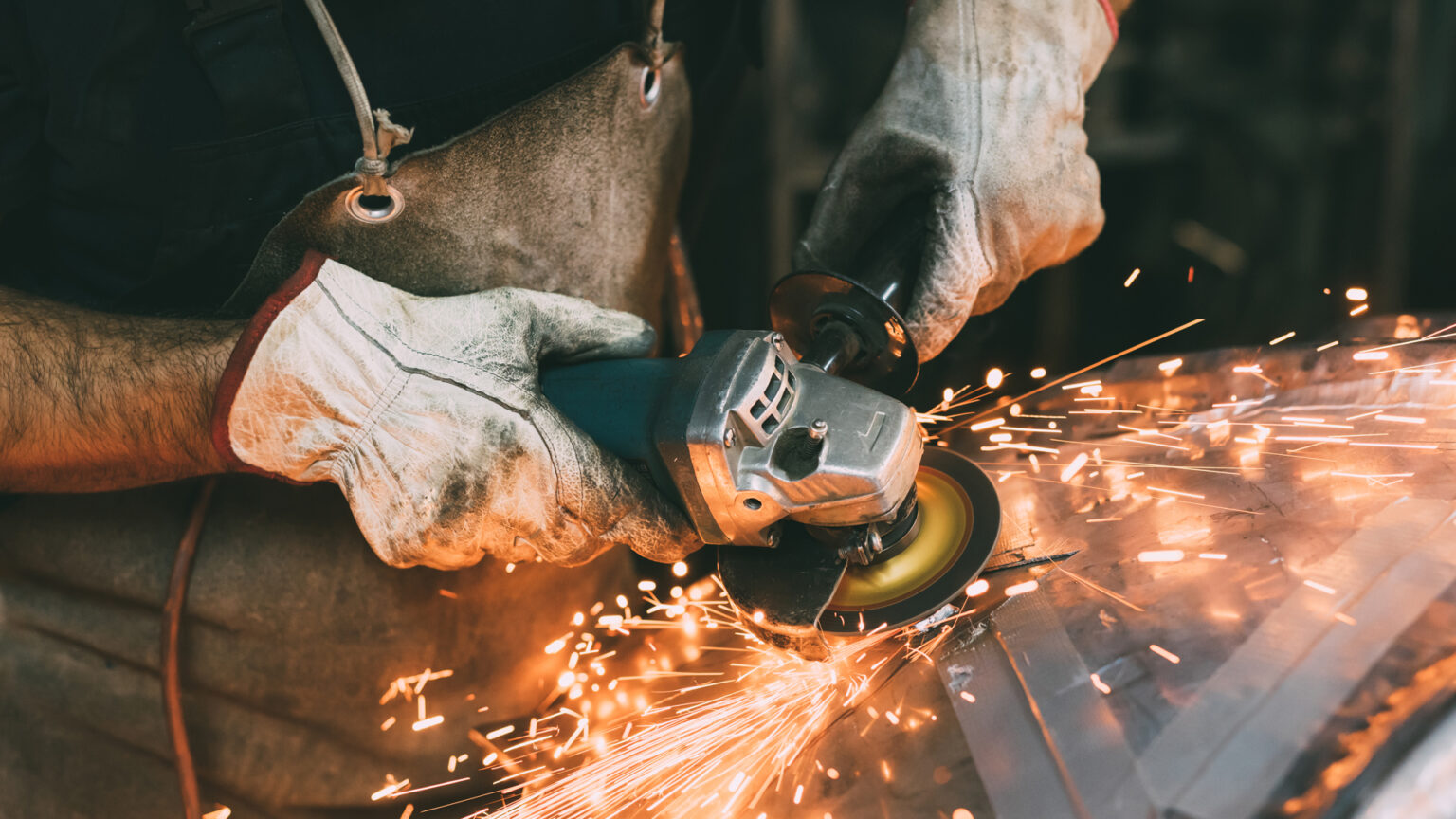 Pracownik w skórzanych rękawicach ochronnych używa szlifierki kątowej, z której lecą iskry podczas obróbki metalu w warsztacie