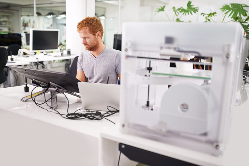 Projektant 3D pracujący przy biurku z tabletem graficznym i laptopem, w tle widoczna nowoczesna drukarka 3D w biurze projektowym.